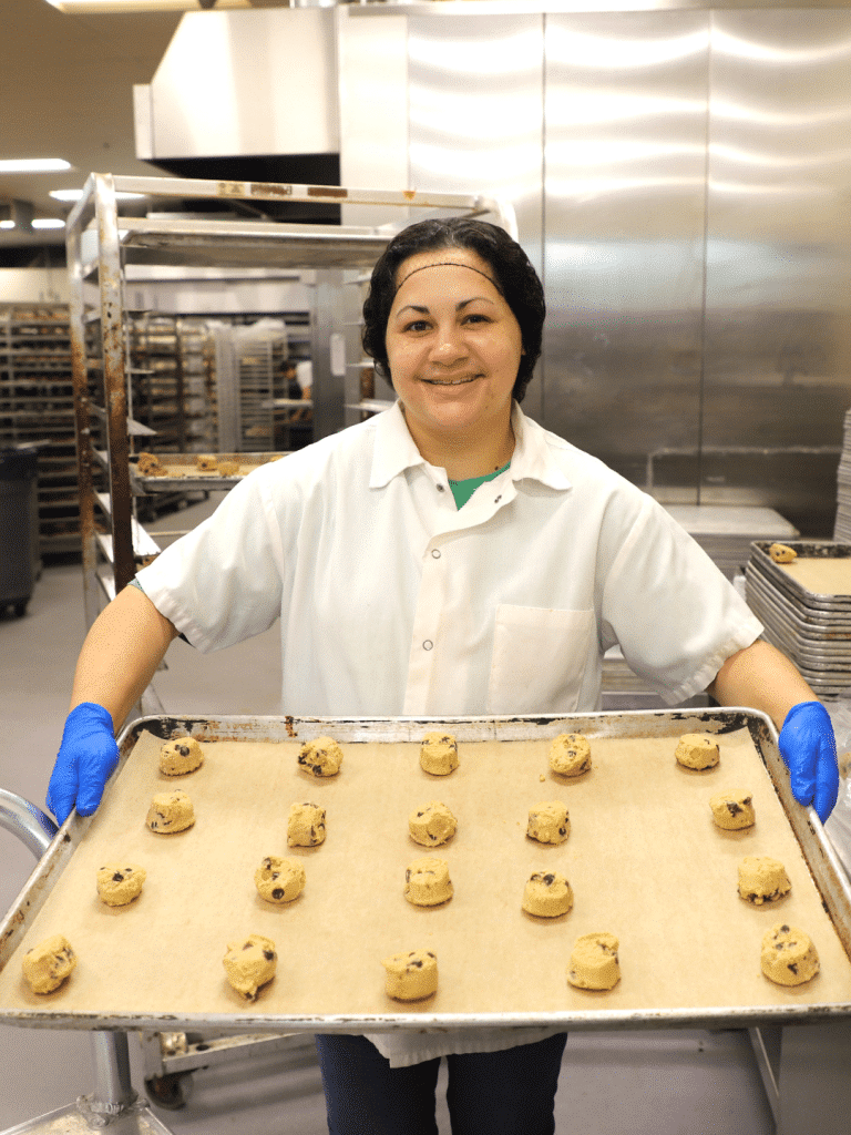 Smiling baker in a white coat and blue gloves holds a tray of raw chocolate chip cookie dough balls in an industrial kitchen, conveying warmth at Busch's Bakehouse in Clinton, MI.