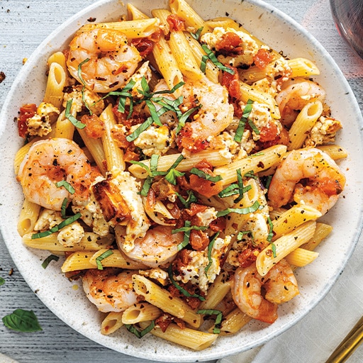 A white bowl filled with penne pasta, shrimp, crumbled feta cheese, and diced tomatoes, garnished with fresh basil and sprinkled with black pepper.