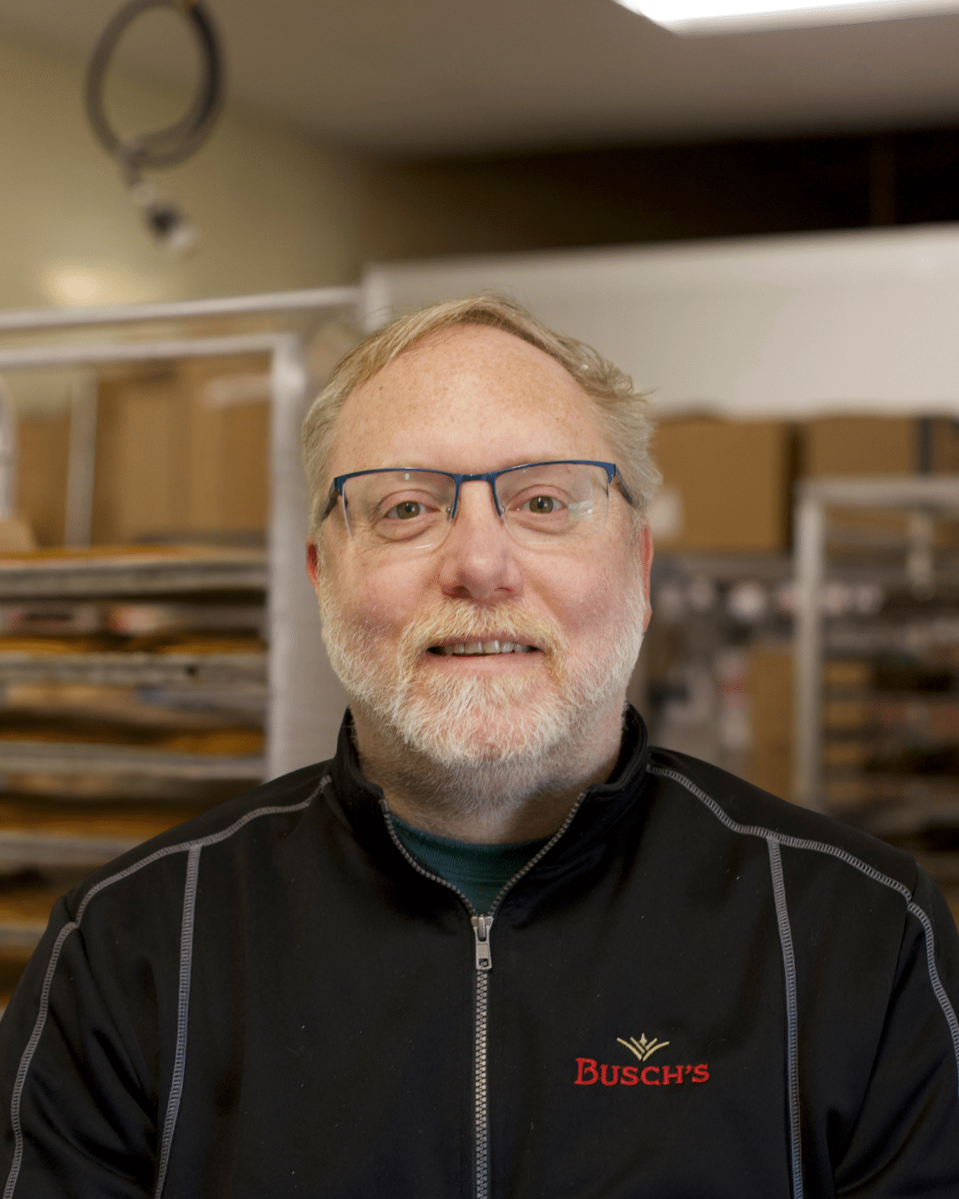 Man with glasses and a beard wearing a black jacket with "Busch's" logo, smiling warmly. Background shows trays in a bakery or kitchen setting. Working in the Busch's Bakehouse in Clinton, Michigan.