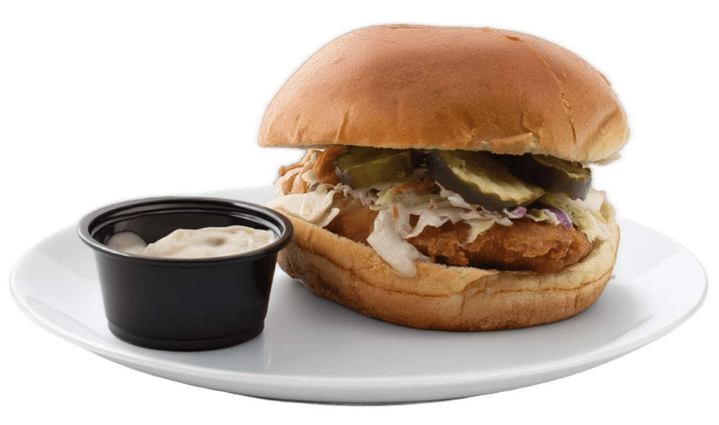 A crispy fish sandwich topped with coleslaw and pickles on a white plate, accompanied by a small black cup of creamy tarter sauce