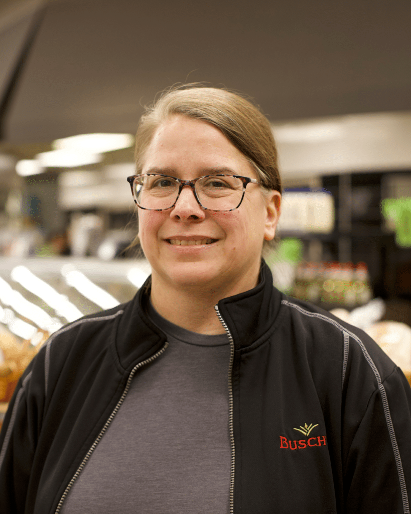 A person in glasses and a black Busch jacket smiles in a grocery store. Bright lights and shelves of products are visible in the background, creating a welcoming atmosphere, who works in Busch's Bakehouse in Clinton, MI.