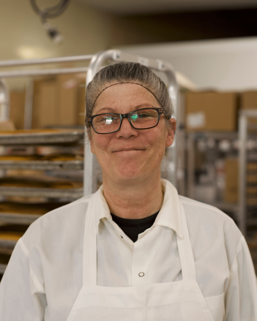 Smiling woman in glasses wearing a white apron and shirt stands in a bakery kitchen. Background shows baking trays and a warm, welcoming atmosphere. Working in the Busch's Bakehouse in Clinton, Michigan.