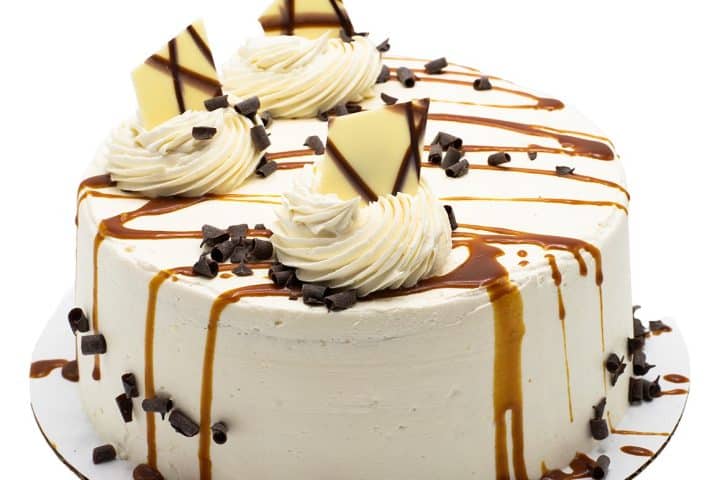 A round vanilla cake topped with swirls of whipped cream, chocolate shavings, and caramel drizzle. Decorative chocolate pieces add elegance. A custom cake from Busch's Fresh Food Market in Clinton, Michigan.