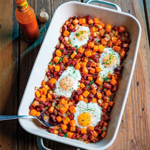 Baked dish with sunny-side-up eggs nestled in seasoned sweet potatoes and sausage cubes, on a wooden table with hot sauce nearby. Cozy and appetizing.