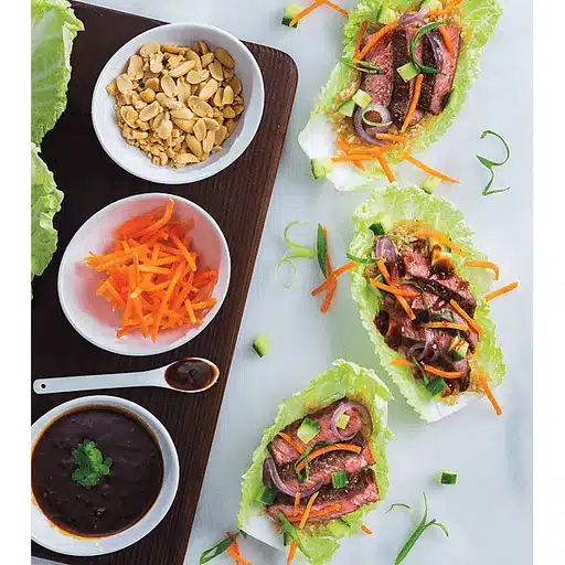 Three lettuce wraps beside bowls of peanuts, shredded carrots, and sauces on a wooden board. The wraps are filled with grilled meat and veggies.