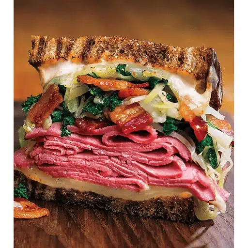 A stacked sandwich with layers of pastrami, melted cheese, lettuce, crispy bacon, and sauerkraut on toasted rye bread, conveying a hearty and savory vibe.