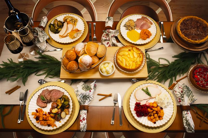 Graphic showing a festive tablescape with plated holiday meals, including turkey, ham, beef, pies, rolls, potatoes, wine, and assorted sides.